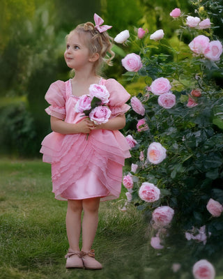 Girl standing next to pink rose flowers wearing faeella blush pink ruffle dress with puff sleeves and pearl neckline perfect for holiday photoshoot