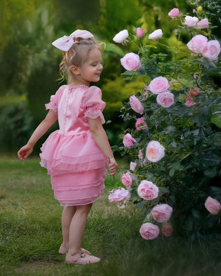 Girl standing next to pink rose flowers with back side wearing faeella blush pink ruffle dress with puff sleeves and pearl neckline perfect for holiday photoshoot
