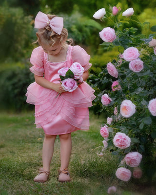 Girl standing next to pink rose flowers wearing faeella blush pink ruffle dress with puff sleeves and pearl neckline perfect for holiday photoshoot