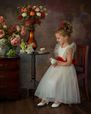 Young girl in a white dress with red accents standing in front of floral arrangements in Faeella's enchanted snow princess gown, perfect for weddings.