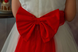 Young girl in a white dress with red accents standing in front of floral arrangements in Faeella's enchanted snow princess gown, perfect for weddings.