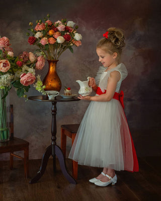 Young girl in a white dress with red accents standing in front of floral arrangements in Faeella's enchanted snow princess gown, perfect for weddings.