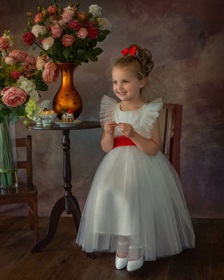 Young girl in a white dress with red accents standing in front of floral arrangements in Faeella's enchanted snow princess gown, perfect for weddings.