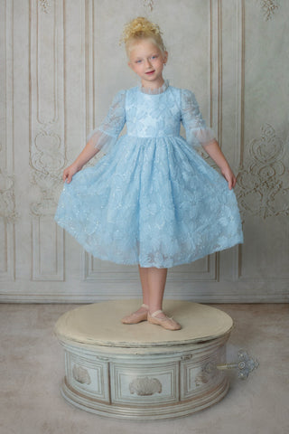 Young girl in a light blue faeella dress, sky petal music box dress standing on a decorative stool against a patterned wall.