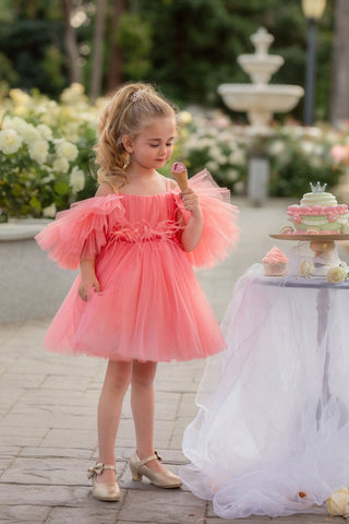 Young girl in a pink dress standing outdoors with a fountain and flowers in the background. Perfect faeella dress for photoshoots and portraits.