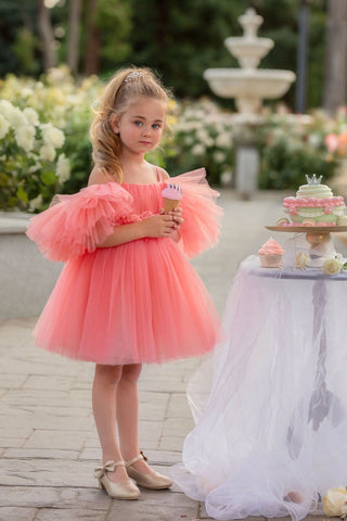 Young girl in a pink dress standing outdoors with a fountain and flowers in the background. Perfect faeella dress for photoshoots and portraits.