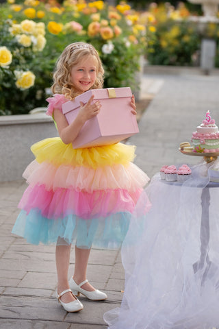 Young girl in a colorful faeella party dress holding a pink gift box outdoors with flowers and a table in the background.
