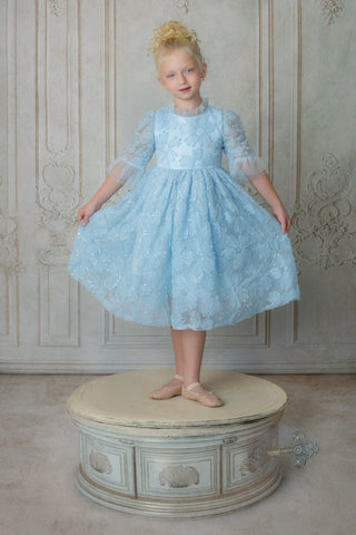 Young girl in a light blue faeella dress, sky petal music box dress standing on a decorative stool against a patterned wall.