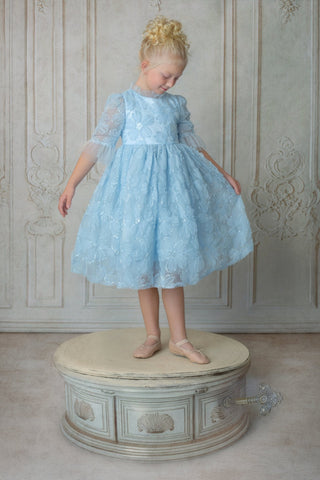 Young girl in a light blue faeella dress, sky petal music box dress standing on a decorative stool against a patterned wall.