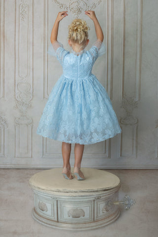 Young girl in a light blue faeella dress, sky petal music box dress back standing on a decorative stool against a patterned wall.